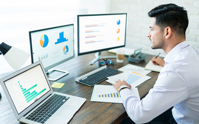 Młody biznesmen przegląda wykresy różnych inwestycji akcji UniCredit na monitorach i kartkach papieru.